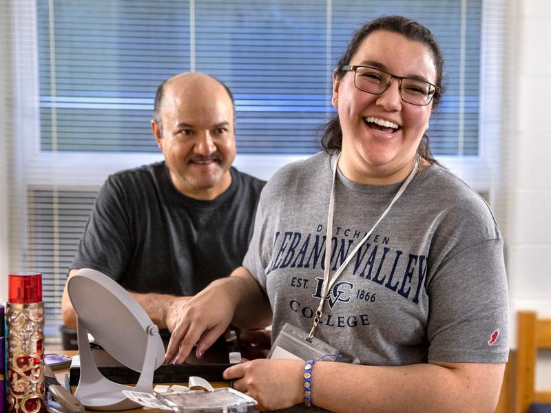 A young woman laughs while arranging items on the top of her dresser. She is wearing glasses and a name badge lanyard around her neck. A man stands behind her, watching and smiling.